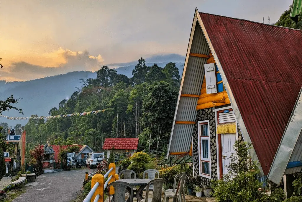 A colorful wooden cabin in a mountainous landscape with lush greenery, surrounded by parked vehicles and a scenic sunset sky